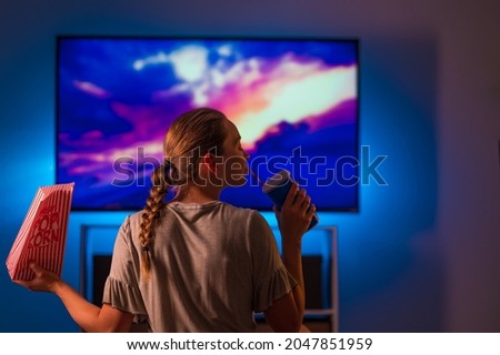 A girl in pajamas sits on an ottoman in front of the TV. She is holding a pack of popcorn and a soda drink. Neon light. Watching your favorite films and programs, fast food, rest, relaxation.