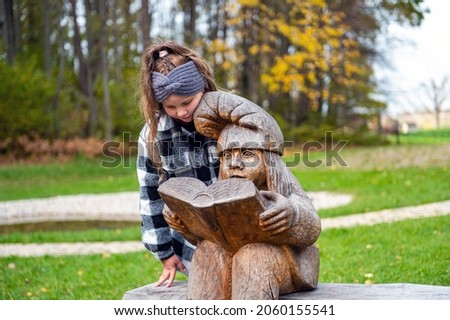 a girl in the park on a bench next to a wooden dwarf sculpture, the dwarf reads a book, Tervete