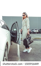 A Girl In Parking Lot Of Shopping Center, Getting Into Car, Opens Door To White Business Sedan Woman In Raincoat, Sneakers, Black Bag In Hands. In Summer And Fall In City