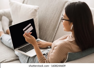 Girl paying online with credit card and laptop, sitting on sofa at home. Computer with blank screen