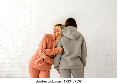 A girl in a peach warm fleece tracksuit smiles with her eyes closed and lies on the shoulder of another girl in a gray fleece suit against a background of white brickwork with space for text.