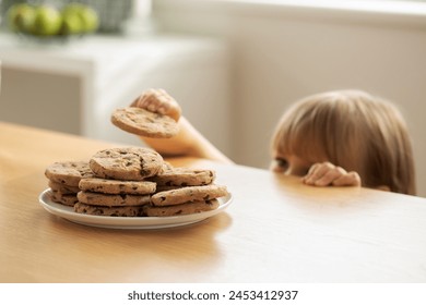 A girl is peeping at an appetizing stack of freshly baked chocolate cookies on the table. The girl hides under the table to secretly eat cookies. - Powered by Shutterstock