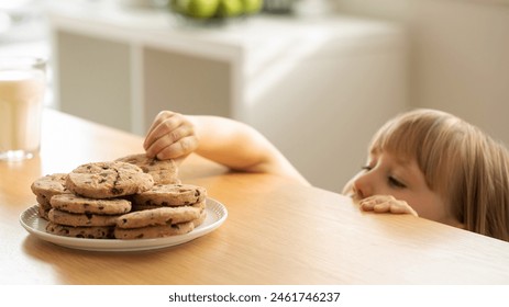 A girl is peeping at an appetizing stack of freshly baked chocolate cookies on the table. The girl hides under the table to secretly eat cookies.