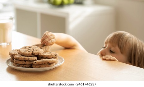 A girl is peeping at an appetizing stack of freshly baked chocolate cookies on the table. The girl hides under the table to secretly eat cookies. - Powered by Shutterstock