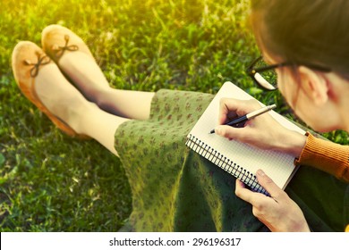 Girl With Pen Writing On Notebook On Grass Outside