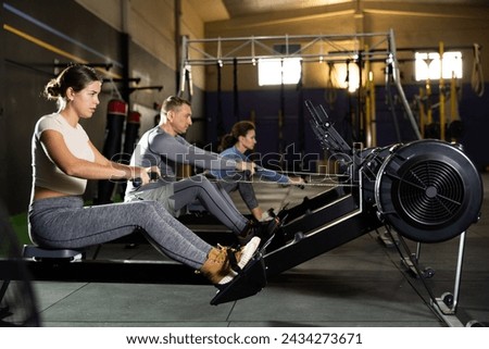 Girl performs exercises with rowing machine. Female gym visitor performs series of repetitions movements on simulator. Shot of sexy muscular woman in sportswear and good physique on grey background