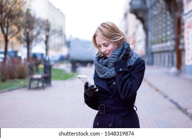 Girl With A Phone In The Autumn Park.  Woman On A Cloudy Day Dressed In Seasonal Weather, Looking At Her Cell Phone. Young Blonde Lady Is Typing A Text Message On Smartphone