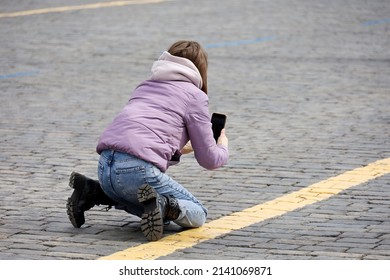 Girl Photographer In Coat And Jeans Taking Pictures On Smartphone Camera Squatting On The Street. Woman Tourist, Female Fashion In Spring