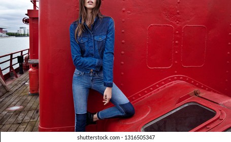 A girl photomodel. She is posing to the camera. blue denim shirt and pant, black boots, red background, wonderful fashion photo