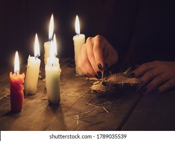 A girl pierces a voodoo doll with needles, close-up. Esoteric and occult rituals on a wooden table in a dark room with many candles. The concept of revenge, causing harm to a rival. Magic background.