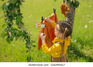 Girl with pigtails hangs washed wet clothes and soft plush toys on clothesline with clothespins under a tree in the garden. Small helper close-up. laundry of children's clothes and drying in fresh air