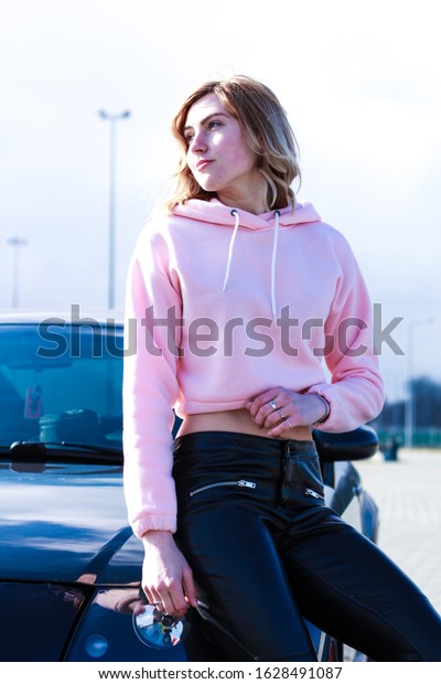 Girl in pink crop\
top. Stylish streetwear for teens. Shooting near a black car. Black\
eco leather pants.