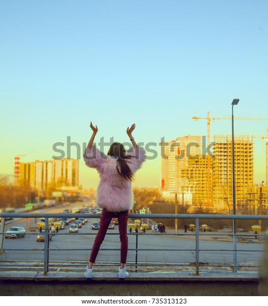 girl in pink fur coat and red leather trousers\
hands up above highway middle. back, rear view of asian woman stand\
against urban road with hard traffic and many modern houses and\
building