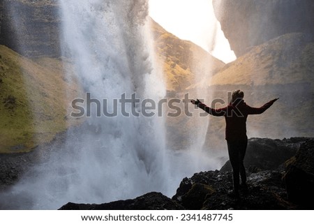 A girl in a pink jacket with arms outstretched standing in a cave on the rocks behind the stunning famous Kvernufoss waterfall, Iceland	