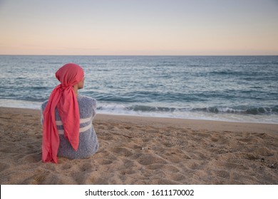 Girl with pink scarf and blue sweater sitting on the shore of the beach at sunset looking at the horizon. Breast cancer concept. - Shutterstock ID 1611170002