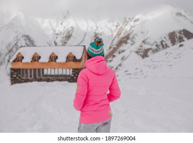 A girl in a pink, warm coat and colorful hat stands with her back to a cozy wooden building with large mountains behind it. Everything around is covered with snow.
