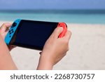 Girl playing game on handheld console on sea background, selective focus on hand. Summer leisure on a beach