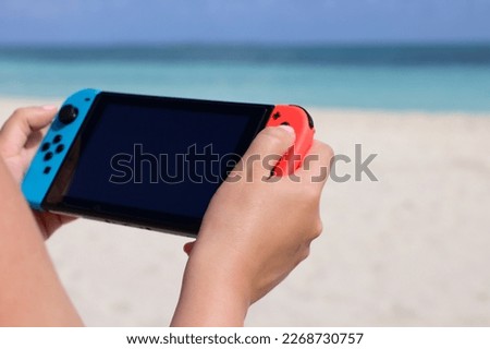 Girl playing game on handheld console on sea background, selective focus on hand. Summer leisure on a beach