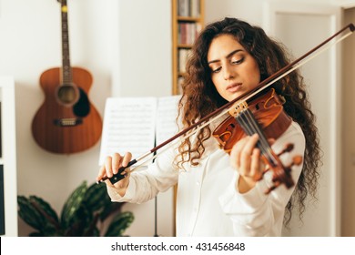 Girl playing violin. Young woman studying music alone at home in the living room with natural and soft light. Curly long and brunette hair, elegant dressed.  - Powered by Shutterstock