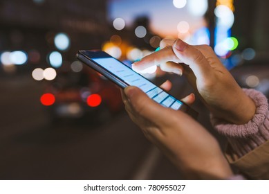 Girl pointing finger on screen smartphone on background illumination bokeh color light in night atmospheric city, using in hands texting mobile phone. Using cellphone at night