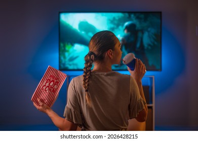 A Girl With Popcorn And A Drink Sits In The Room And Watches TV. Night Time. Insomnia, Watching Your Favorite Movie, Loneliness, Leisure. Shooting From The Back.