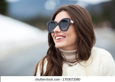 Girl posing on road on winter woods background. Glamorous funny young woman with smile wearing stylish sweater, white fashionable fur coat and sunglasses. Fur and fashion concept. Beautiful people.