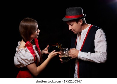 Girl Pouring Beer From A Keg Into A Mug Of Guy In Octoberfest. Couple In Nationa Ethnic Dress Having Fun With Alcohol. Girlfriend And Boyfriend Posing In Studio With Black Background In Oktoberfest