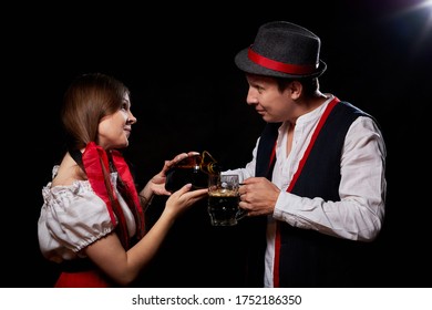 Girl Pouring Beer From A Keg Into A Mug Of Guy In Octoberfest. Couple In Nationa Ethnic Dress Having Fun With Alcohol. Girlfriend And Boyfriend Posing In Studio With Black Background In Oktoberfest