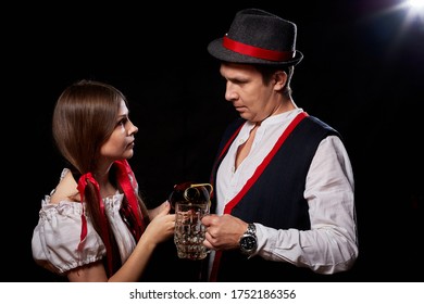 Girl Pouring Beer From A Keg Into A Mug Of Guy In Octoberfest. Couple In Nationa Ethnic Dress Having Fun With Alcohol. Girlfriend And Boyfriend Posing In Studio With Black Background In Oktoberfest