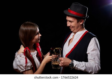 Girl Pouring Beer From A Keg Into A Mug Of Guy In Octoberfest. Couple In Nationa Ethnic Dress Having Fun With Alcohol. Girlfriend And Boyfriend Posing In Studio With Black Background In Oktoberfest