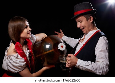 Girl Pouring Beer From A Keg Into A Mug Of Guy In Octoberfest. Couple In Nationa Ethnic Dress Having Fun With Alcohol. Girlfriend And Boyfriend Posing In Studio With Black Background In Oktoberfest