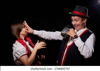 Girl Pouring Beer From A Keg Into A Mug Of Guy In Octoberfest. Couple In Nationa Ethnic Dress Having Fun With Alcohol. Girlfriend And Boyfriend Posing In Studio With Black Background In Oktoberfest