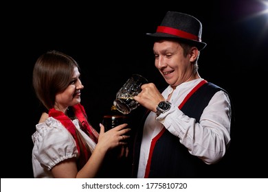 Girl Pouring Beer From A Keg Into A Mug Of Guy In Octoberfest. Couple In Nationa Ethnic Dress Having Fun With Alcohol. Girlfriend And Boyfriend Posing In Studio With Black Background In Oktoberfest