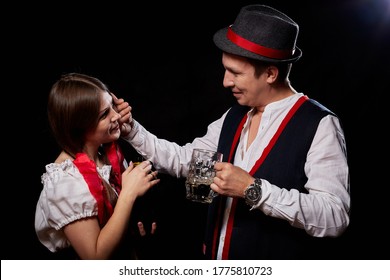 Girl Pouring Beer From A Keg Into A Mug Of Guy In Octoberfest. Couple In Nationa Ethnic Dress Having Fun With Alcohol. Girlfriend And Boyfriend Posing In Studio With Black Background In Oktoberfest