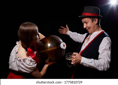 Girl Pouring Beer From A Keg Into A Mug Of Guy In Octoberfest. Couple In Nationa Ethnic Dress Having Fun With Alcohol. Girlfriend And Boyfriend Posing In Studio With Black Background In Oktoberfest