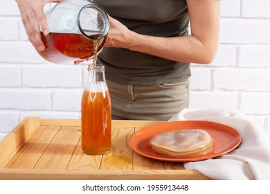 Girl pouring ready homemade kombucha tea into a glass bottle from big jar. Orange plate with scoby. Light bricked background. Healthy fermented beverage.