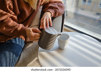 Girl Pours Hot Tea Into A Cup From A Teapot. Enjoy The Comforts Of Home. White Cup Close-up In The Hands Of A Woman In The Rays Of The Sun From The Window. A Cup Of Tea In The Hands Of A Woman