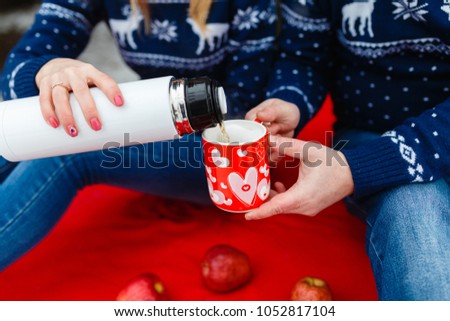 Girl pours tea in a mug with a picture of the heart. Young couples in sweaters sit on red blanket. Wedding picnic in the fresh air in the winter in the forest or mountains. Offer to marry on a picnic.