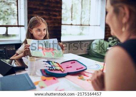 Girl presenting her artwork teacher. Woman assisting schoolgirl during classes at primary school. Child drawing picture sitting at desk in classroom. Girl drawing pictures. Learning at primary school