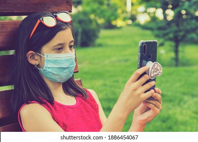 girl in protective mask holding a smartphone. teen girl siting on bench in green park with mobile phone. First stage of loosening coronavirus restrictions and self-isolation. toned