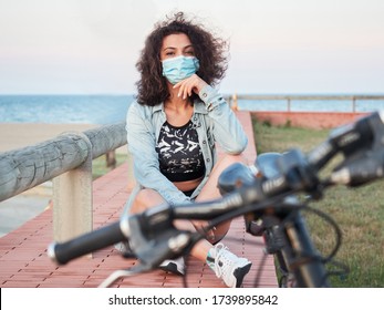 Girl with a protective mask resting in a park after a bike ride. Beach in the background. - Powered by Shutterstock