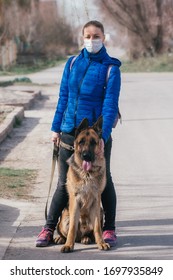 A Girl In A Protective Medical Mask Walks A Dog On The Street. Leisure With A Pet During Quarantine. Walk With A German Shepherd In The Fresh Air. Self-isolation And Protection Mode