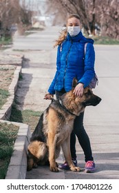 A Girl In A Protective Medical Mask Walks A Dog On The Street. Leisure With A Pet During Quarantine. Walk With A German Shepherd In The Fresh Air. Self-isolation And Protection Mode