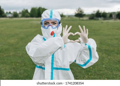 A girl in a protective suit ppe from the covid-19 virus. On the face is a medical mask and safety glasses. On hands rubber gloves. Holds a heart. Sign language. Corona virus. Quarantine. Pandemic.  - Powered by Shutterstock