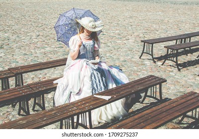 a girl in a puffy dress of the 18th century in a beautiful hat and in the evening reading a book sitting on a bench - Shutterstock ID 1709275237