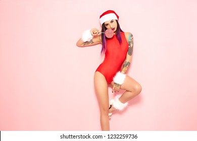 Girl With Purple Hair Tips And Tattoo On Her Arm Dressed In Red Swimsuit, Santa's Hat And White Fur Bracelets Holds A Lollipop At Her Face On The Background Of Pink Wall