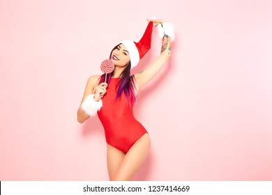 Girl With Purple Hair Tips And Tattoos On Her Arm Dressed In Red Swimsuit, Santa's Hat And White Fur Bracelets Holds A Lollipop And Raises Up Hats Top On The Background Of Pink Wall