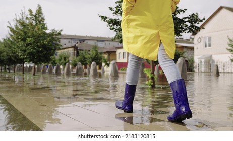 Girl In Raincoats Rubber Boots Walk Along Road Flooded With Torrential Rains. Their Feet Walk Through Puddles City. Splashing Water Sides, Flood Street. Puddle Water Splash Concept. Summer Rainfall