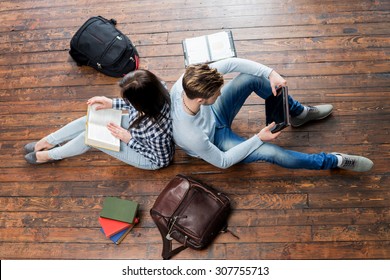 Girl Reading A Book And Boy Using A Tablet Leaning On Each Other On Wooden Floor Having Notebooks And Bags Around Them.  