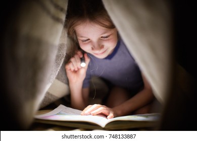 girl is reading a book under a blanket with a flashlight in a dark room at night